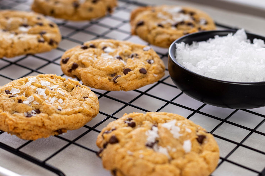 Chocolate chip cookies topped with flaky finishing salt beside a bowl of coarse sea salt on a cooling rack.